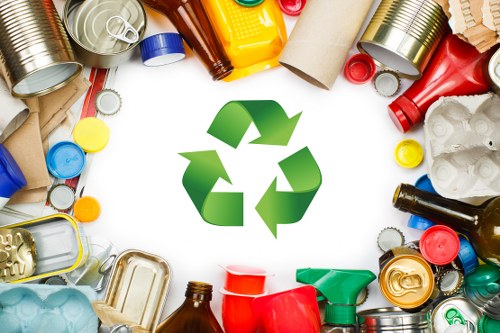 A wide circular arrangement of various discarded household items including aluminium cans, glass bottles, plastic containers, cardboard tubes, and crumpled paper, surrounding a green recycling symbol centered in the image. The items exhibit a range of colours such as red, yellow, blue, brown, and metallic silver, and vary in texture from smooth plastic to reflective metal and rough cardboard. The background is plain and white, with the scattered waste objects creating a contrast that emphasizes their forms. The collection of rubbish exemplifies typical waste materials that a professional rubbish removal service like Office Clearance Enfield might collect from commercial or residential properties within areas like Enfield postcodes. The scene is evenly lit, with no shadows or external environmental context provided, focusing solely on the assortment of waste items and the recycling icon, visually representing the importance of waste segregation and disposal in the context of environmentally conscious rubbish services in the Enfield locality.