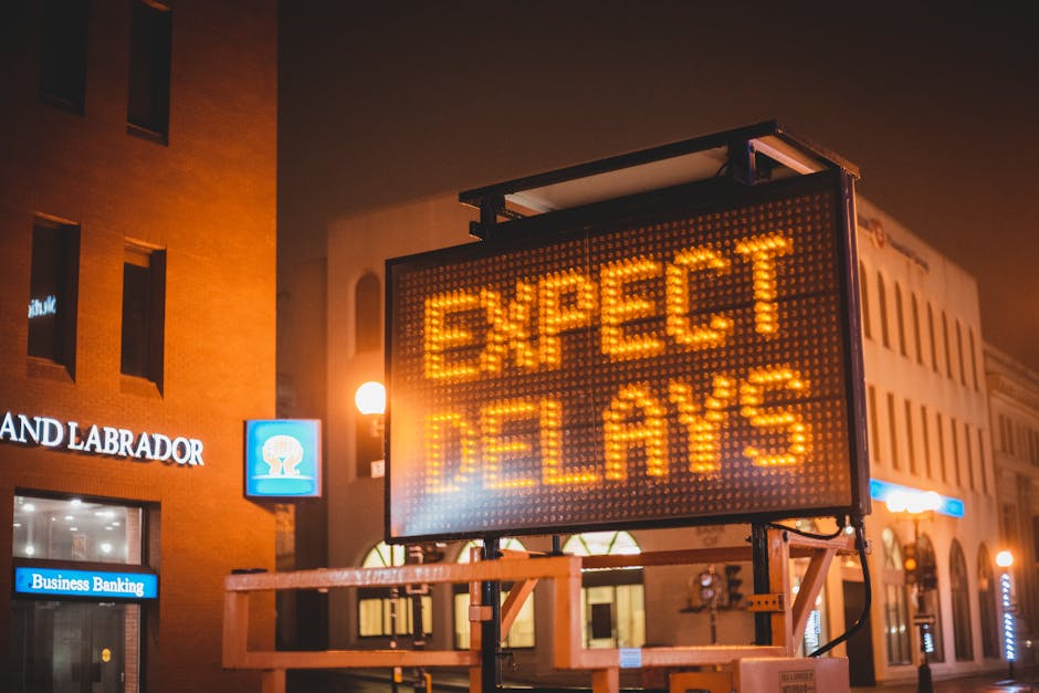 A large electronic road sign displaying the message 'EXPECT DELAYS' in bright amber pixels is mounted on a metal support structure on a city street at night. The sign is illuminated, casting a glow on