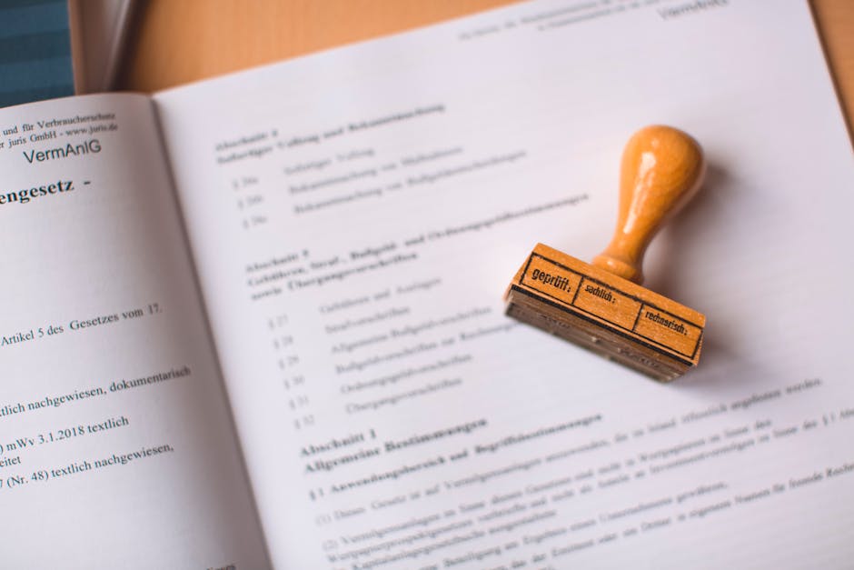 Close-up of a person wearing a dark blazer and a green shirt, holding a pen in their right hand and a clipboard in their left hand. The person's hand features a silver ring on the ring finger. The sce