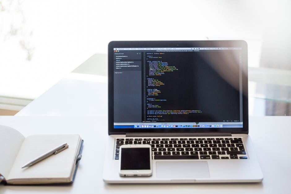 A silver laptop with a black keyboard displaying a dark-themed code editor on its screen, placed on a white desk in a bright environment. To the left of the laptop, there is an open white notebook wit